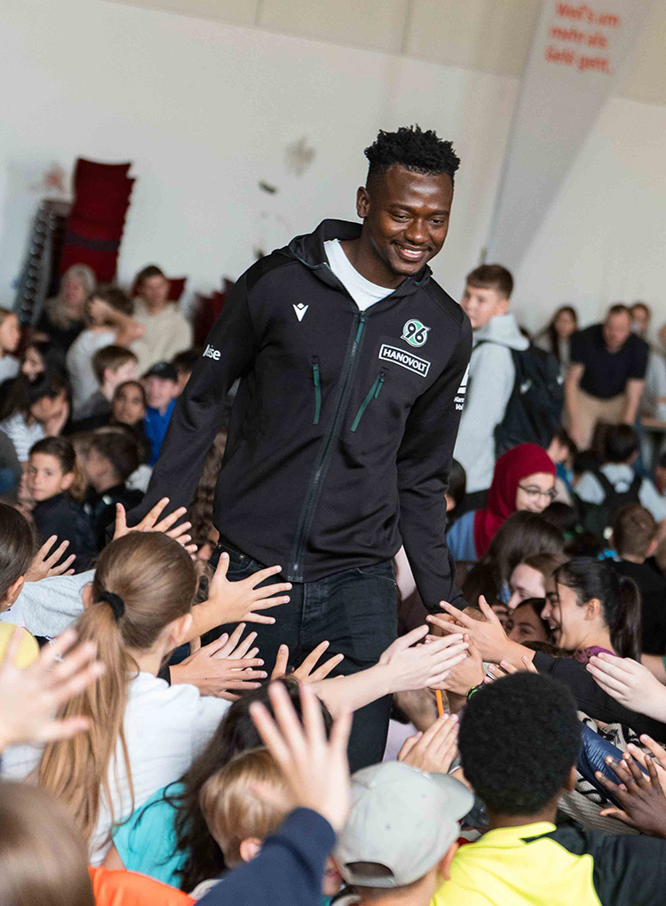 Der 96-Spieler Mustapha Bundu läuft in einer schwarzen 96-Jacke durch ein Pulk von Kindern und klatscht mit ihnen an. 