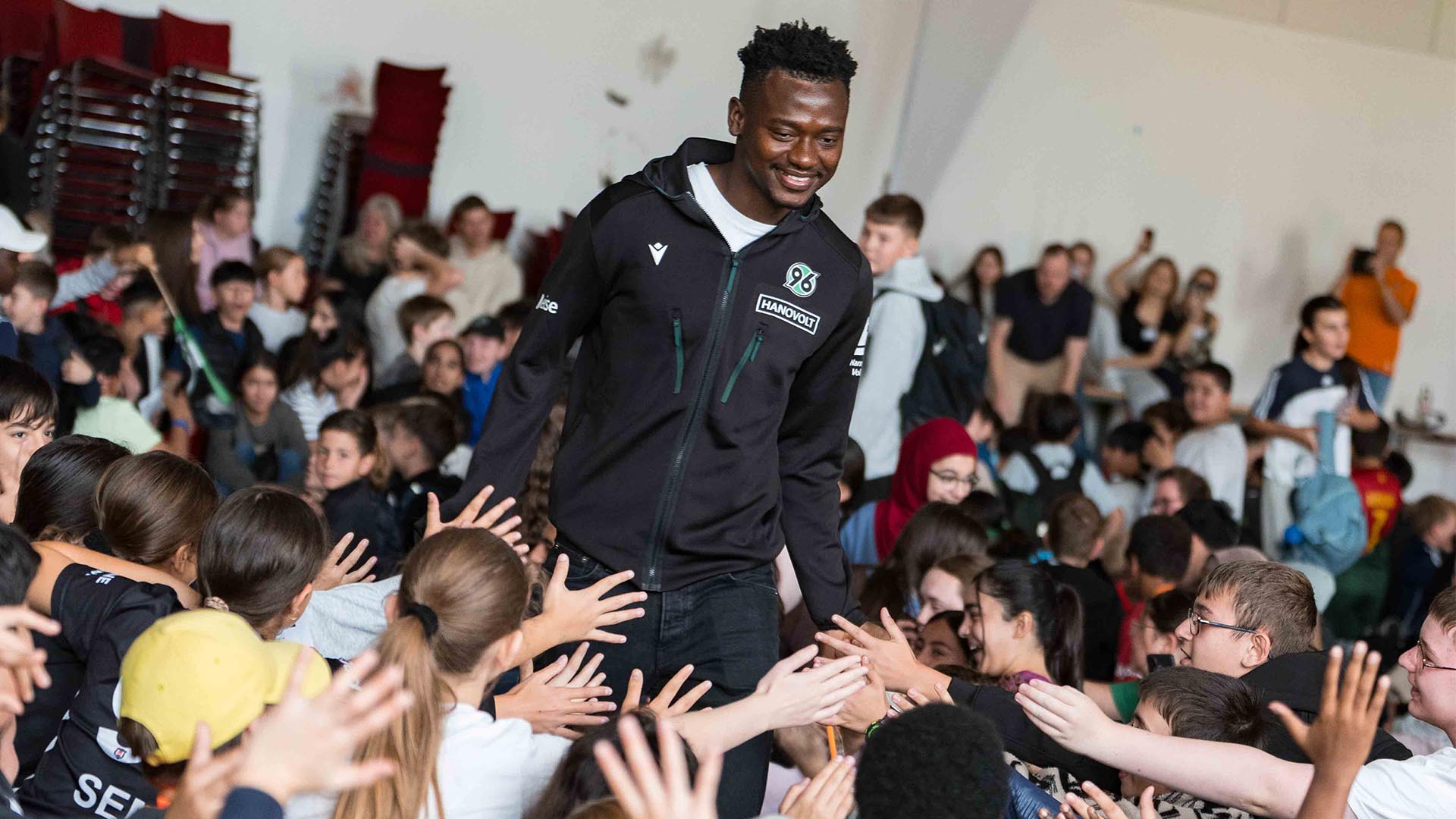 Der 96-Spieler Mustapha Bundu läuft in einer schwarzen 96-Jacke durch ein Pulk von Kindern und klatscht mit ihnen an. 