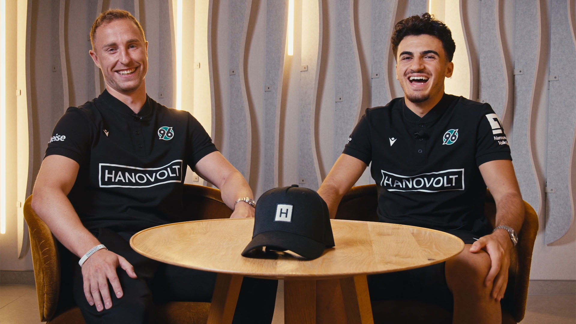 Die 96-Spieler Maurice Neubauer und Husseyn Chakroun sitzen auf Stühlen, vor ihnen ein Holztisch, auf dem eine schwarze Cap liegt. Beide lachen herzlich.