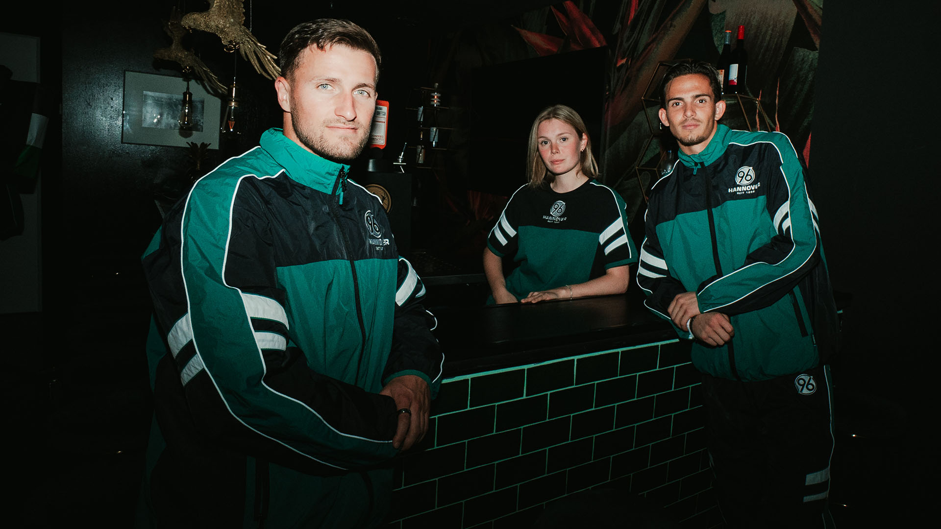 Enzo Leopold, ein weibliches Model und Nahuel Noll präsentieren die Retro-Kollektion an eine Bar gelehnt.