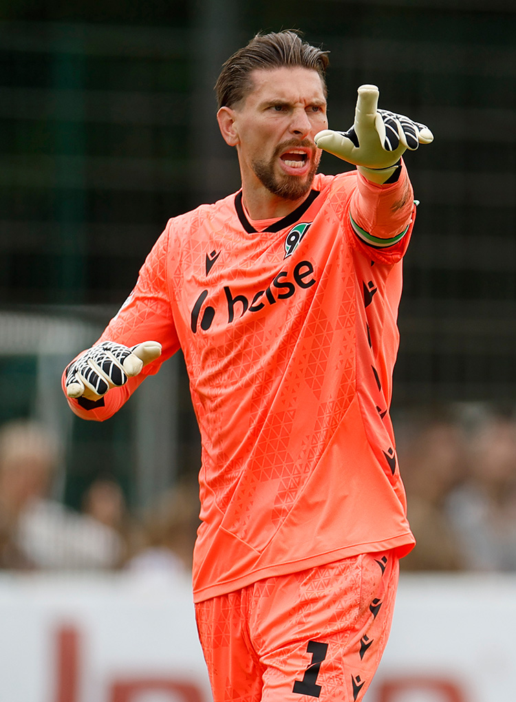 Ron-Robert Zieler im orangefarbenen Torwartrikot ruft etwas und zeigt mit dem linken Arm.