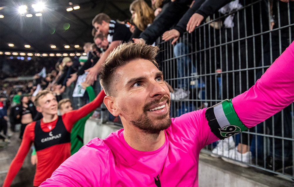 Ron-Robert Zieler im pinken Torwarttrikot klatscht mit den Fans in der Nordkurve ab und strahlt dabei. 
