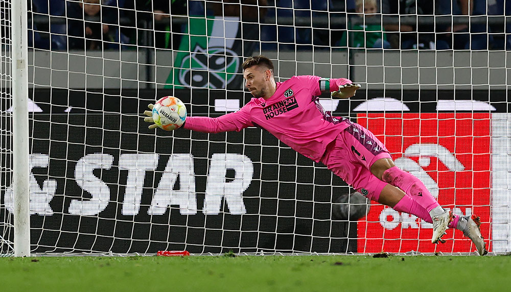 Ron-Robert Zieler im pinken Torwarttrikot springt nach rechts und wehrt einen Ball ab.