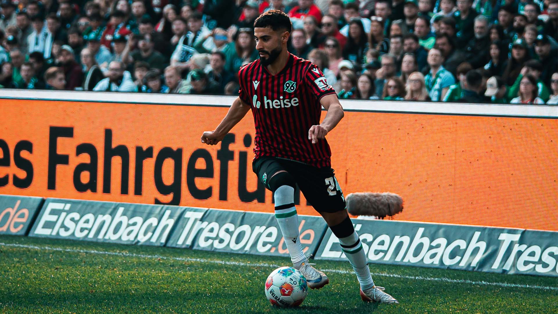 Elias Saad führt an der Seitenlinie mit seinem rechten Außenrist den Ball. Er trägt das rot-schwarz gestreifte Heimtrikot. Im unscharfen Hintergrund sind Zuschauer zu sehen.