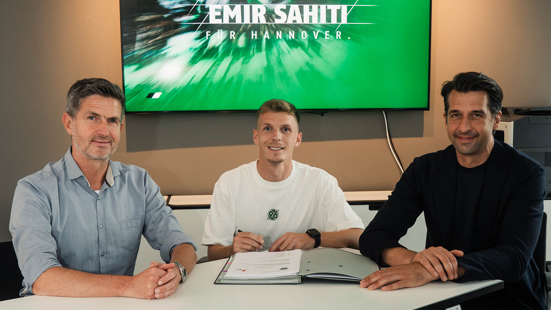 Der neue 96-Spieler Emir Sahiti sitzt in der Mitte, rechts daneben 96-Geschäftsführer Jonas Boldt, links daneben 96-Sportdirektor Ralf Becker, im Hintergrund ein Bildschirm mit Sahitis Namen.