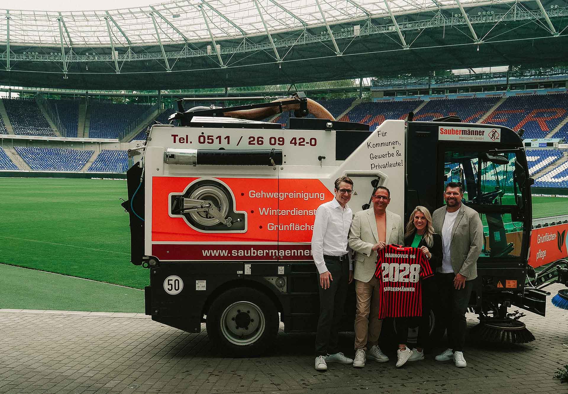 Vier Verantwortliche von Hannover 96, Sportfive und den Saubermännern stehen vor einem Reinigungswagen der Saubermänner im leeren Stadion und halten gemeinsam ein Trikot. 