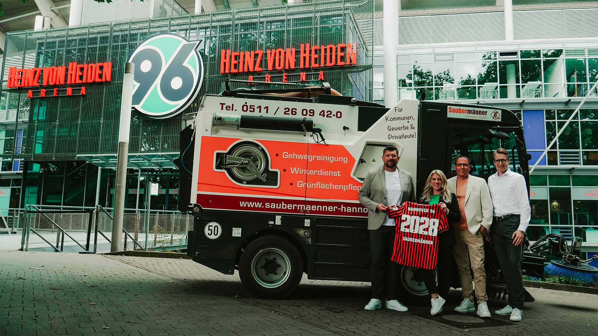 Vier Verantwortliche von Hannover 96, Sportfive und den Saubermännern stehen vor einem Reinigungswagen der Saubermänner vor dem Stadion und halten gemeinsam ein Trikot. 