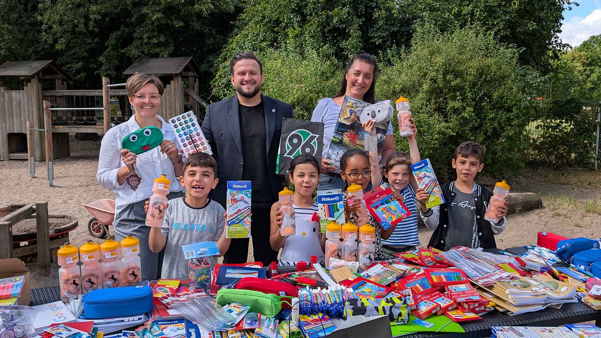 Ein Hannover-96-Mitarbeiter, zwei Mitarbeiterinnen des Familienzentrums St. Franziskus und fünf Kinder jubeln über die Schulsachenspende.
