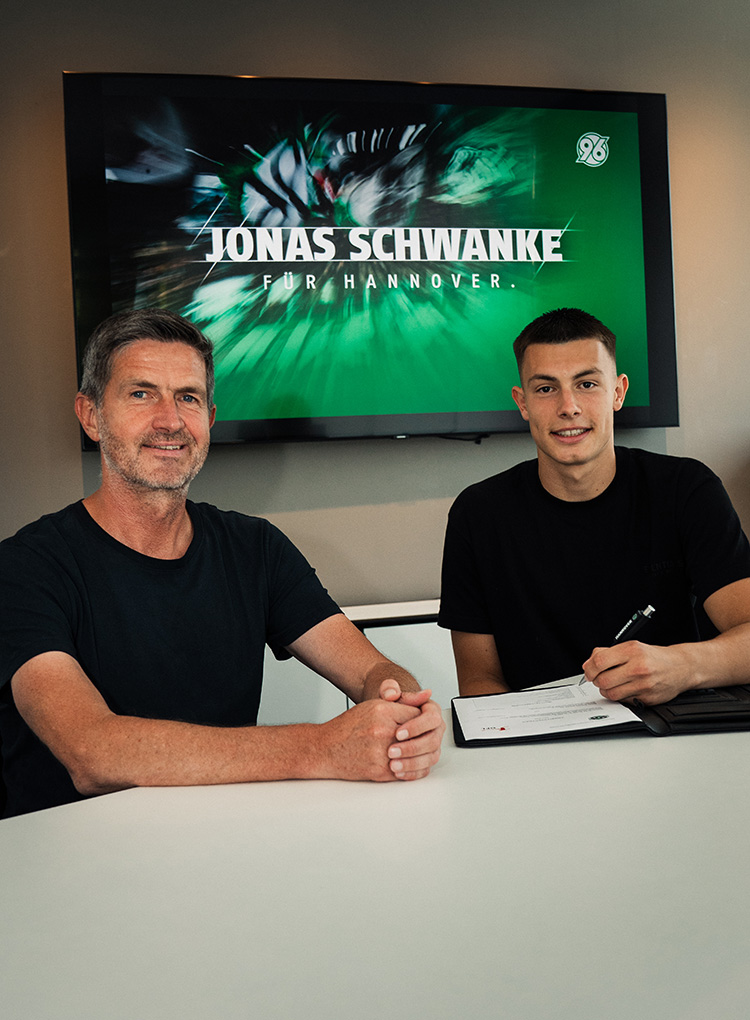 96-Sportdirektor Ralf Becker und Torwart Jonas Schwanke sitzen am Tisch in einem Büro, im Hintergrund steht auf einem Bildschirm "Jonas Schwanke für Hannover".