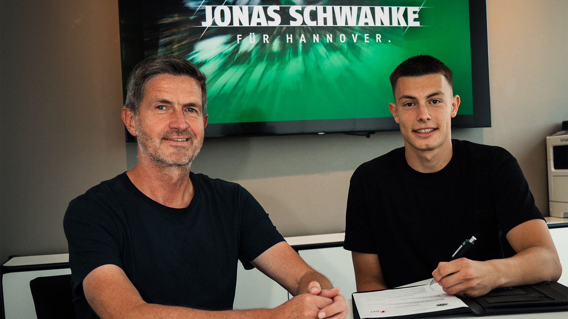 96-Sportdirektor Ralf Becker und Torwart Jonas Schwanke sitzen am Tisch in einem Büro, im Hintergrund steht auf einem Bildschirm "Jonas Schwanke für Hannover".