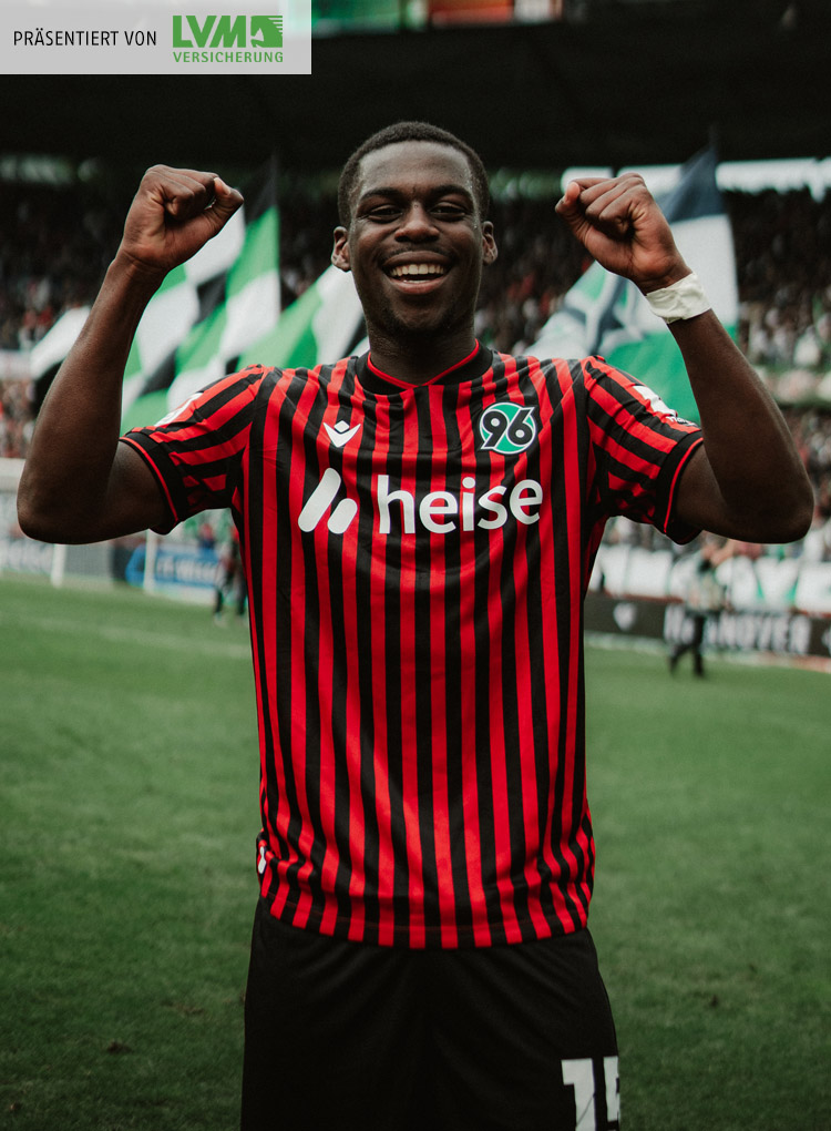 Noel Aseko ballt die Fäuste und jubelt in die Kamera, im Hintergrund sieht man unscharf das volle Stadion und Fahnen.