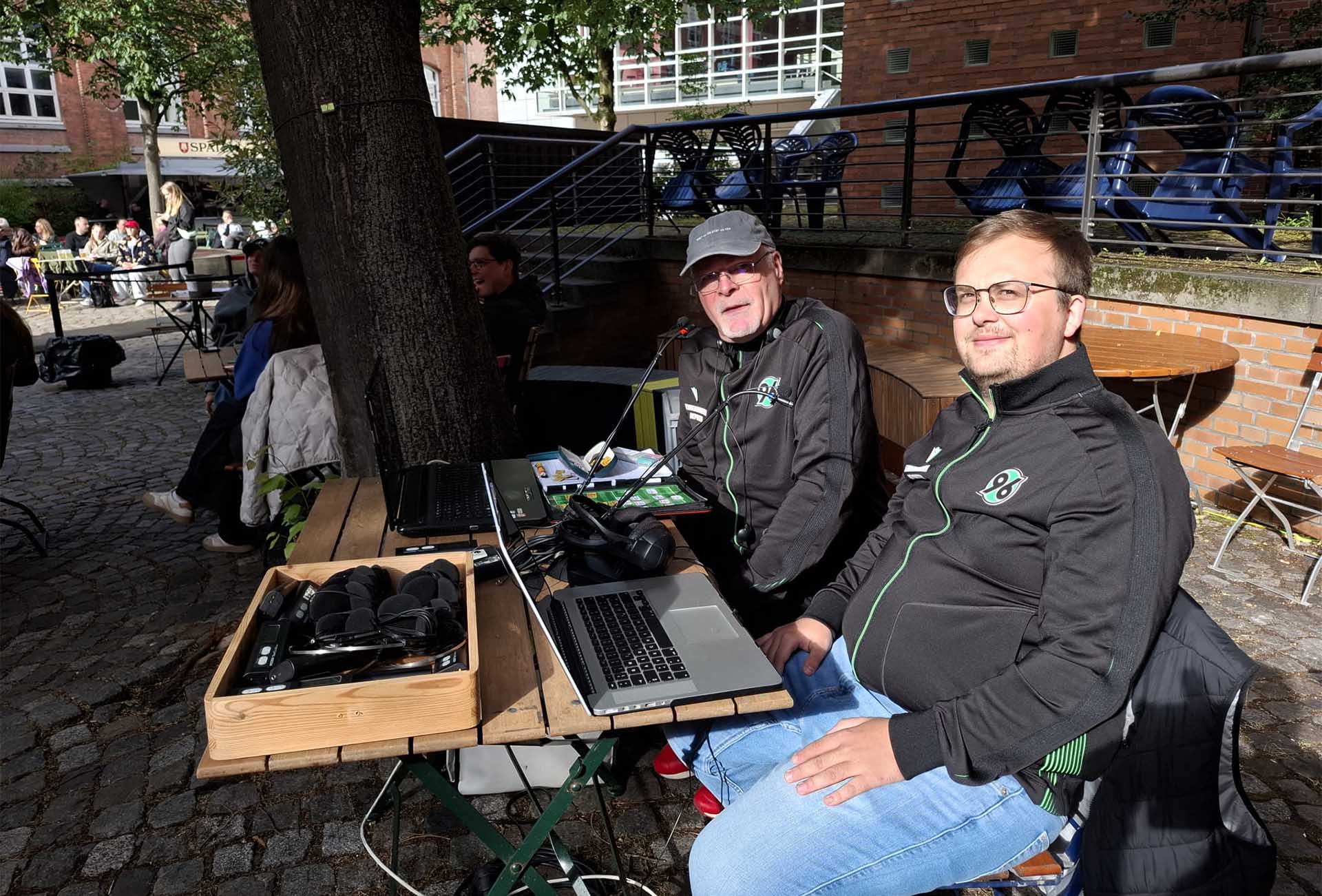 Zwei Männer sitzen im Freien an einem Tisch mit Laptops und Kopfhörern, im Hintergrund sind andere Menschen zu sehen.