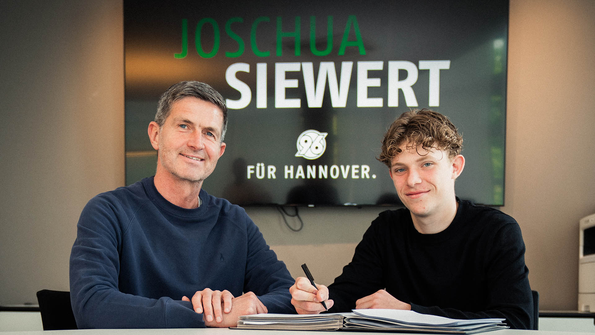 Joshua Siewert mit Ralf Becker bei der Unterschrift, im Hintergrund ein Bildschirm, auf dem groß sein Name steht.