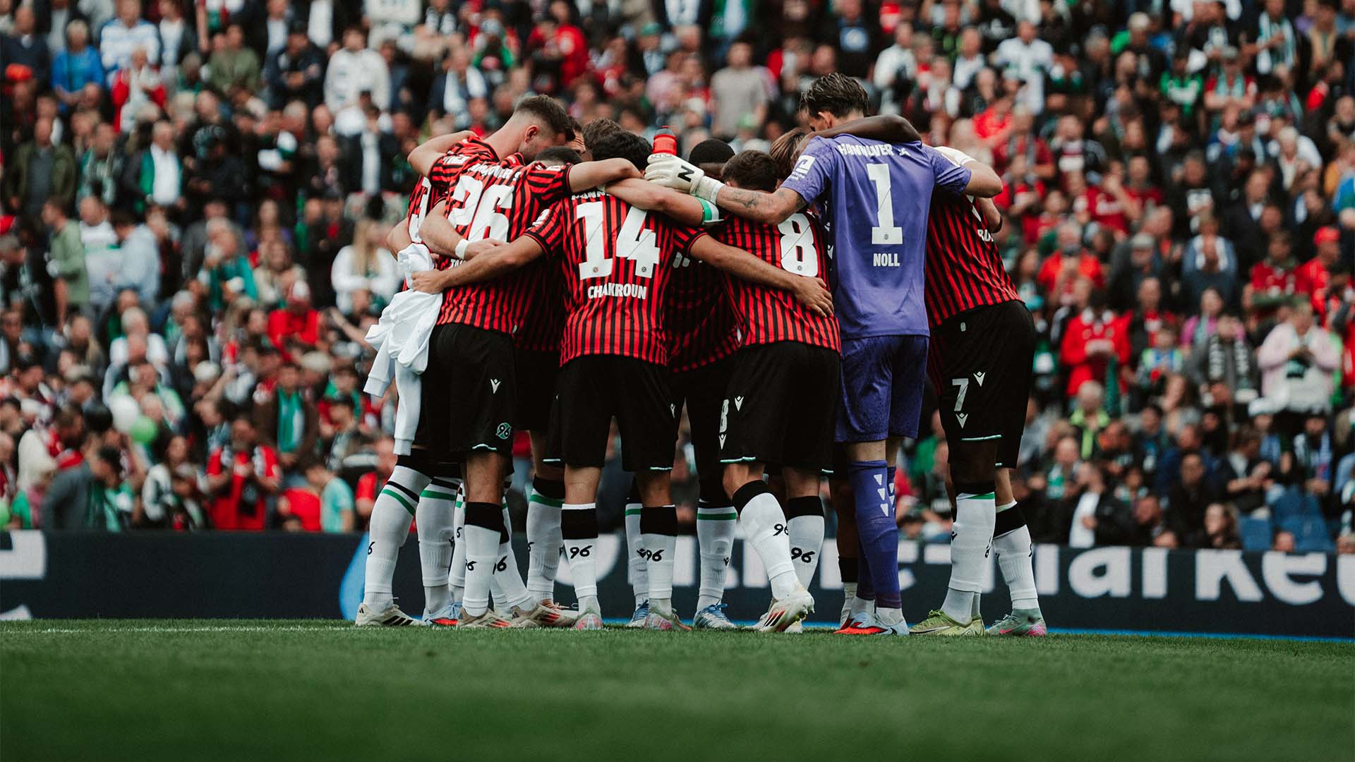 Ein Mannschaftskreis im Stadion, im Hintergrund unscharf Fans auf den Rängen.