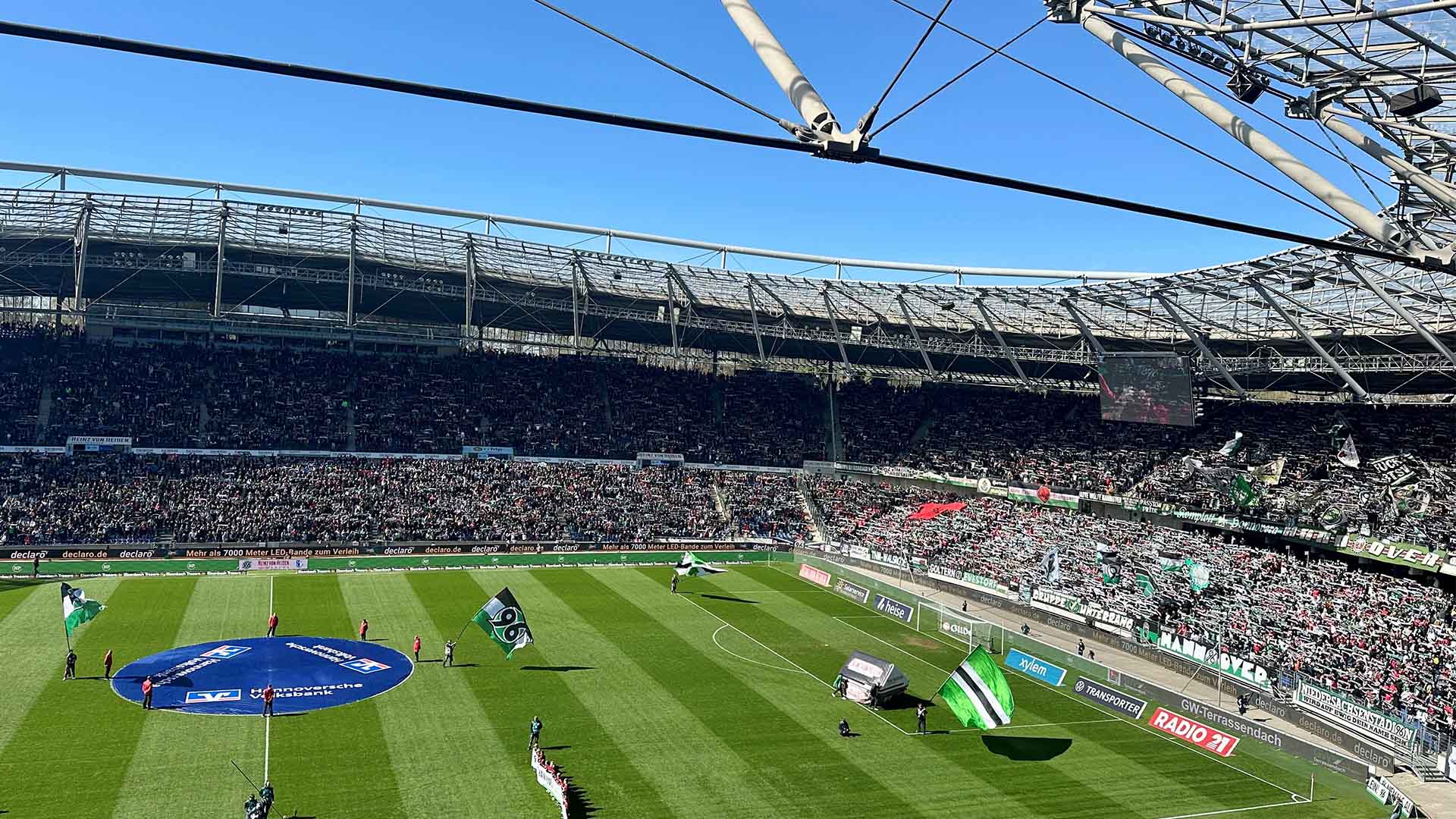 Blick auf die volle Nordkurve der Heinz von Heiden Arena und Teile der Westkurve, auf dem Platz stehen Fahnenschwenker.