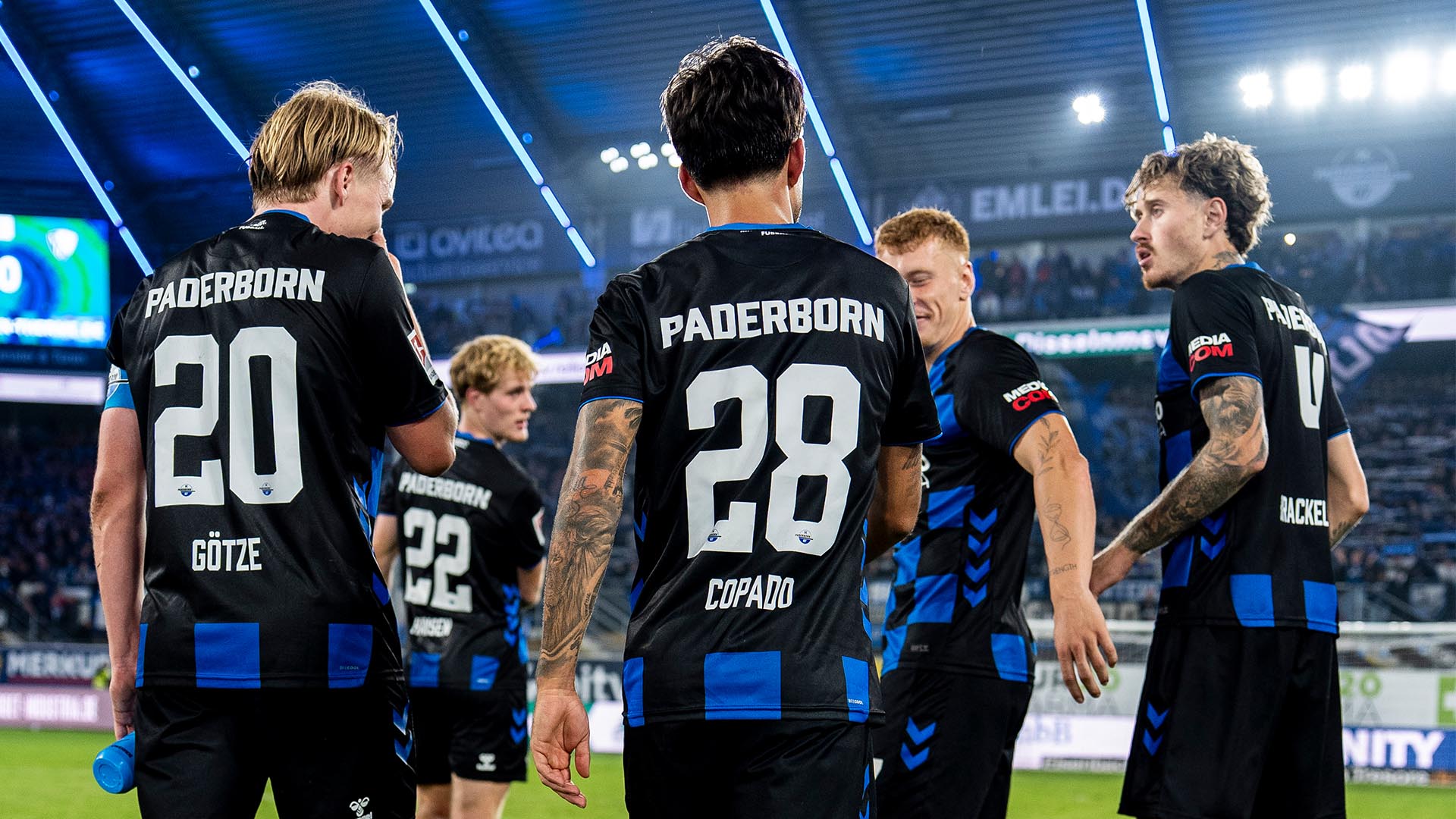Fünf Spieler des SC Paderborn stehen auf dem Rasen zusammen und sprechen miteinander. Im Stadion leuchten die Flutlichter und im Hintergrund sind Zuschauer zu sehen.