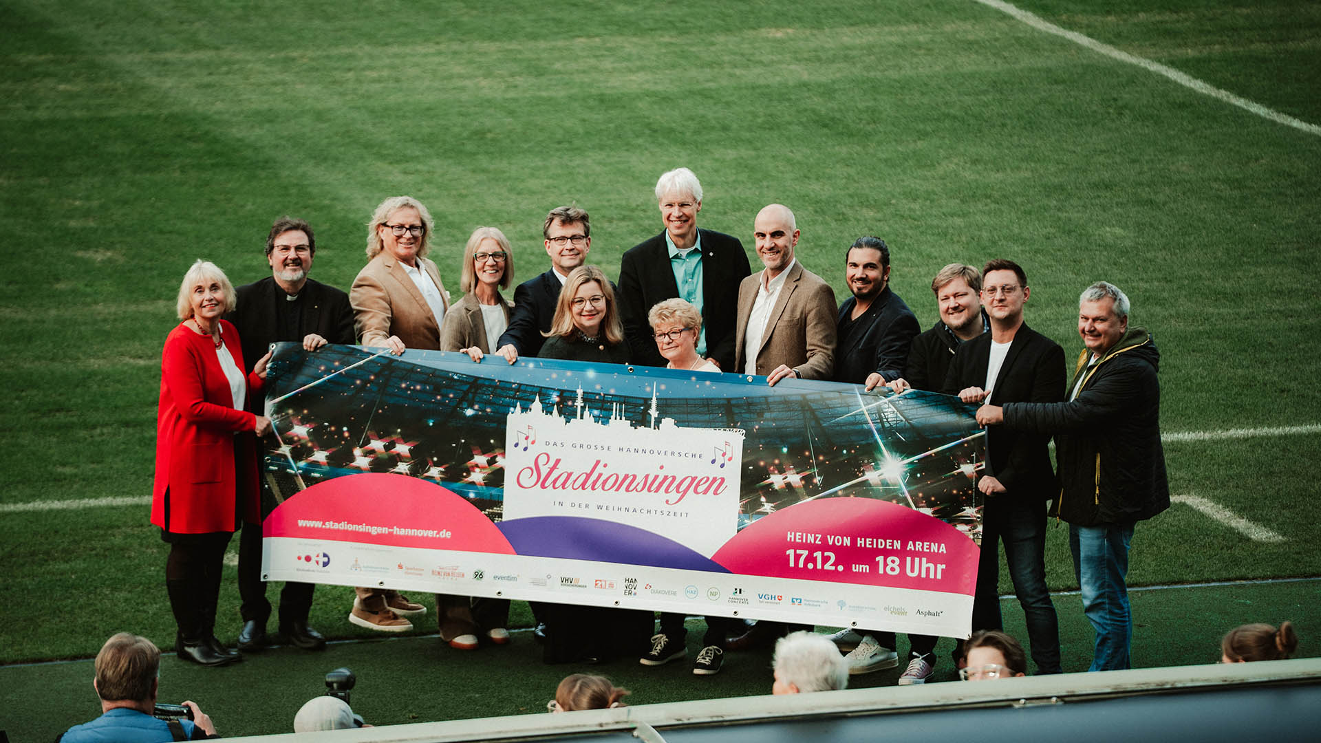 Mehrere Personen halten das Banner für das Stadionsingen hoch. Die Gruppe steht auf dem Rasen der Heinz von Heiden Arena.