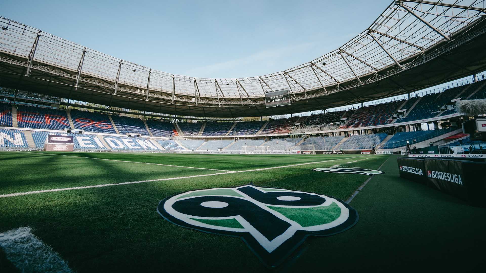 Die fast leere Heinz von Heiden Arena bei Sonnenschein. Auf der Videowall steht "Herzlich willkommen", im Vordergrund liegt ein 96-Logo-Teppich.