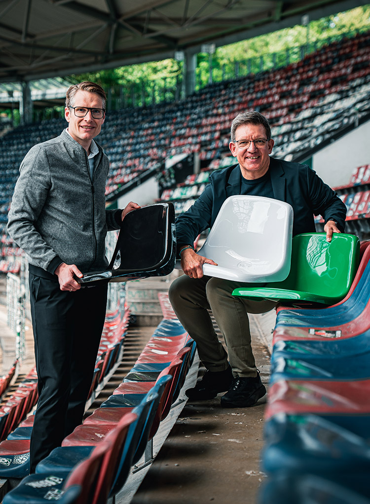 Zwei Männer sitzen auf der Tribüne im Stadion, sie halten drei neue Sitzschalen in Schwarz, Weiß und Grün.
