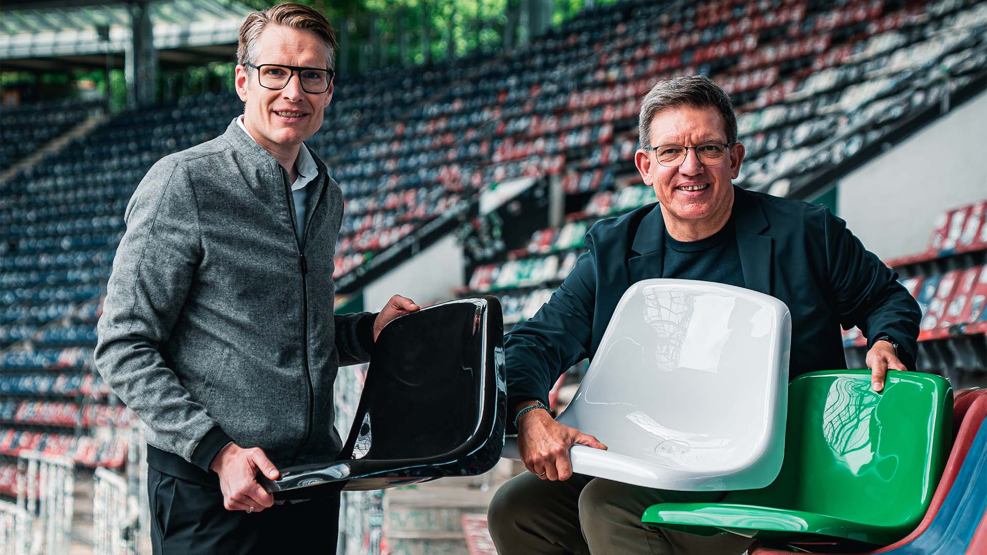Zwei Männer sitzen auf der Tribüne im Stadion, sie halten drei neue Sitzschalen in Schwarz, Weiß und Grün.