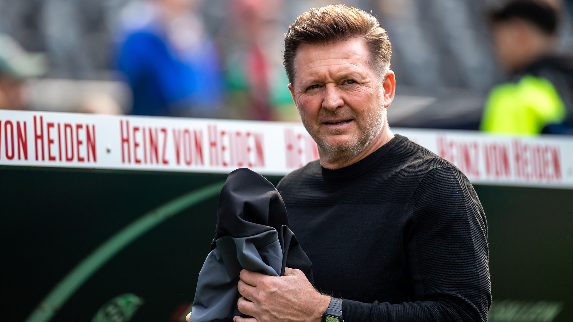 Chistian Titz mit einer Jacke in der Hand im Stadion.