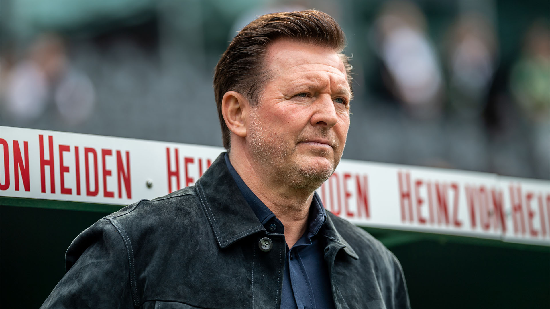 Christian Titz steht vor der Bank im Stadion und schaut leicht fokussiert. 