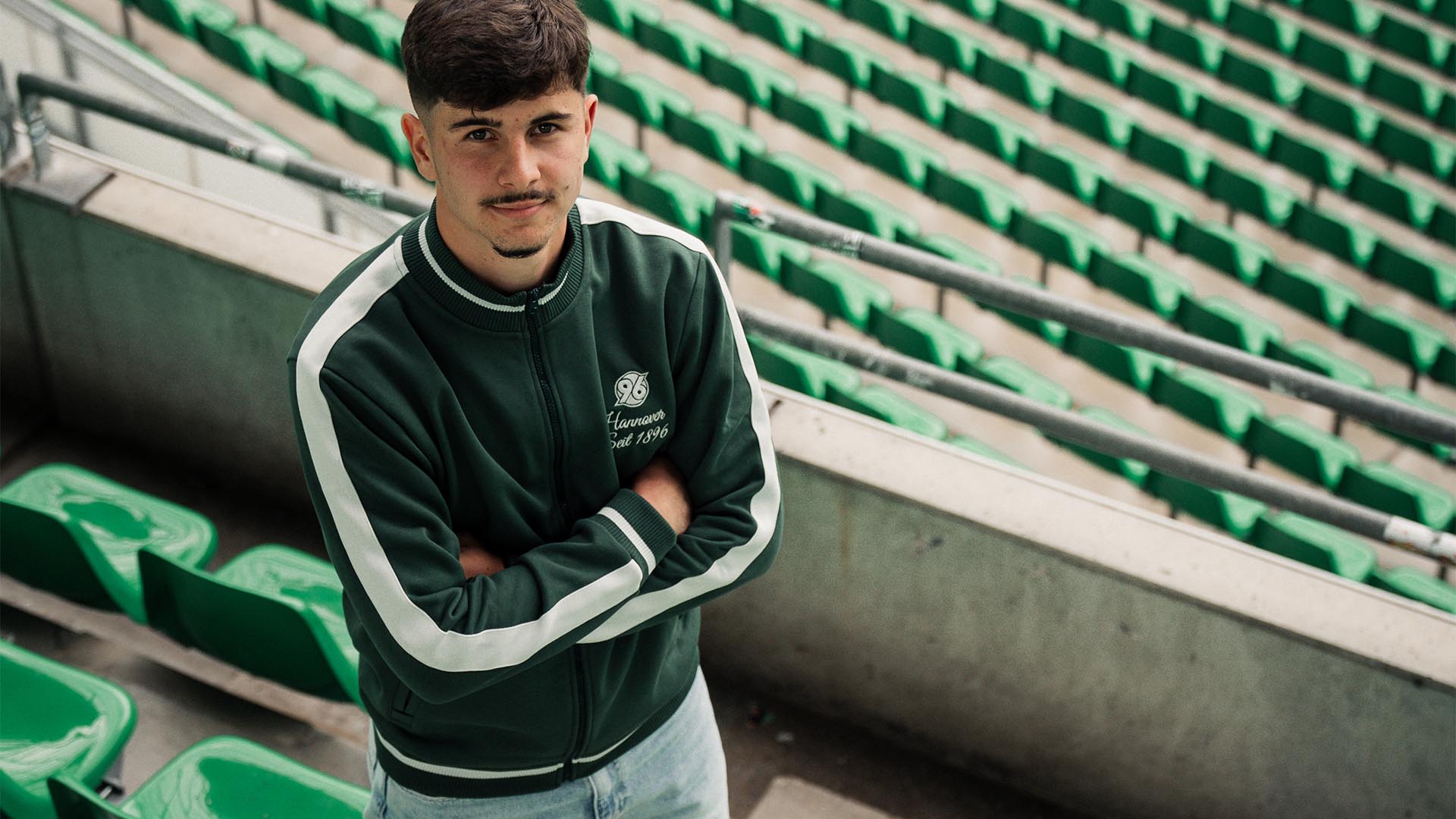 Der neue 96-Spieler Karl Steinmann steht in einem 96-Hoodie auf den Rängen des 96-Stadions und schaut nach oben in die Kamera.
