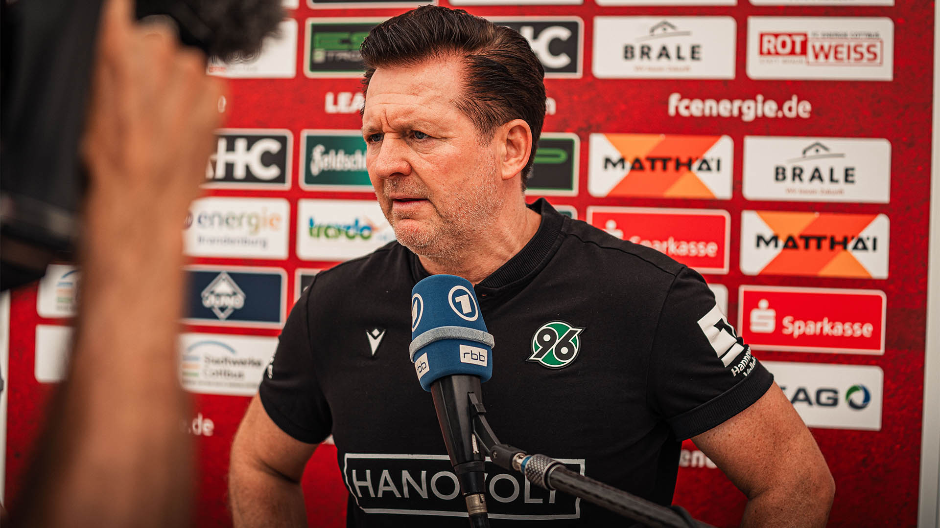 Christian Titz steht vor einer Sponsorenwand und spricht in ein rbb-Mikrofon.