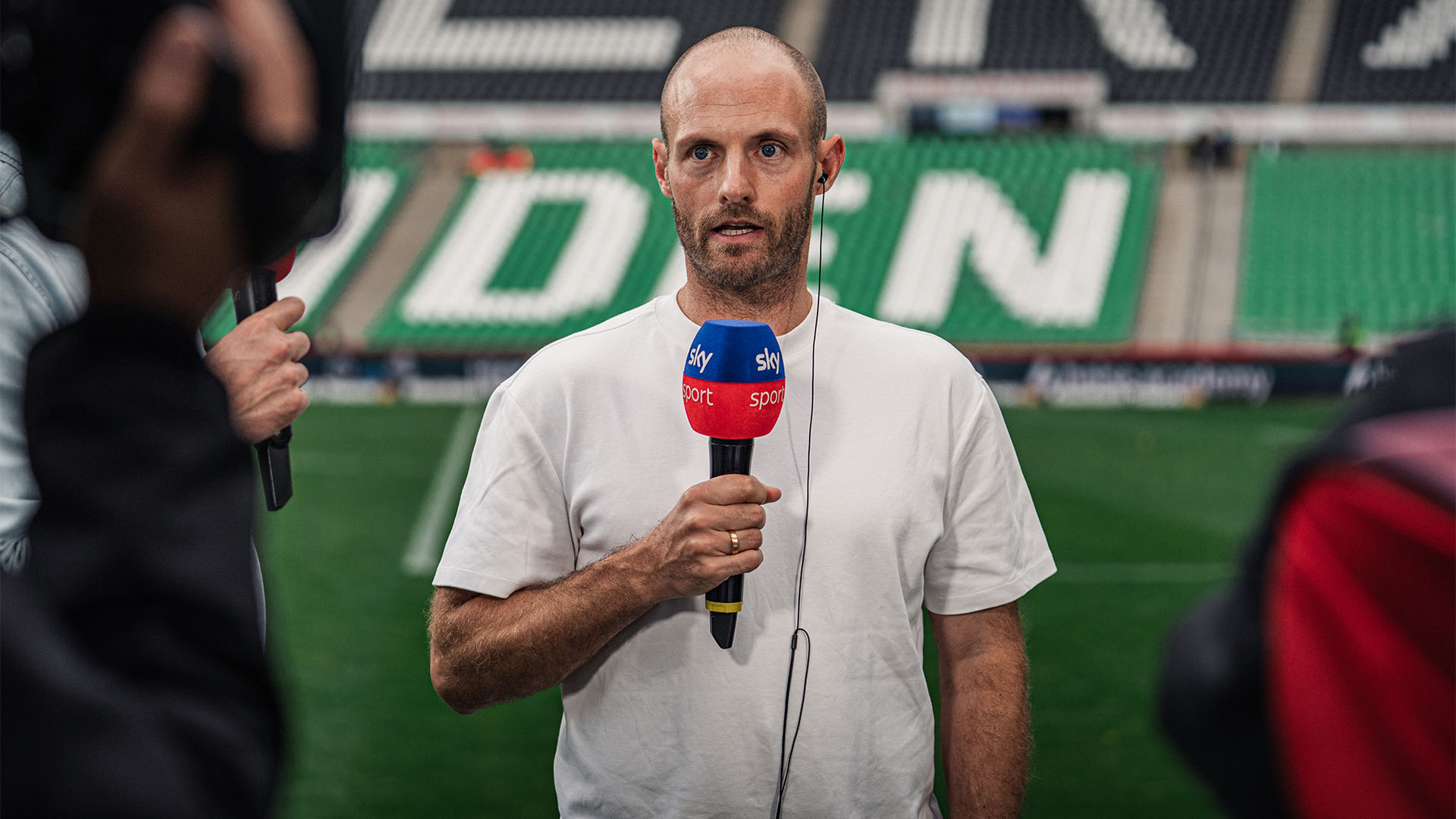 96-Trainer Lars Barlemann im weißen T-Shirt am Sky-Mikrofon.