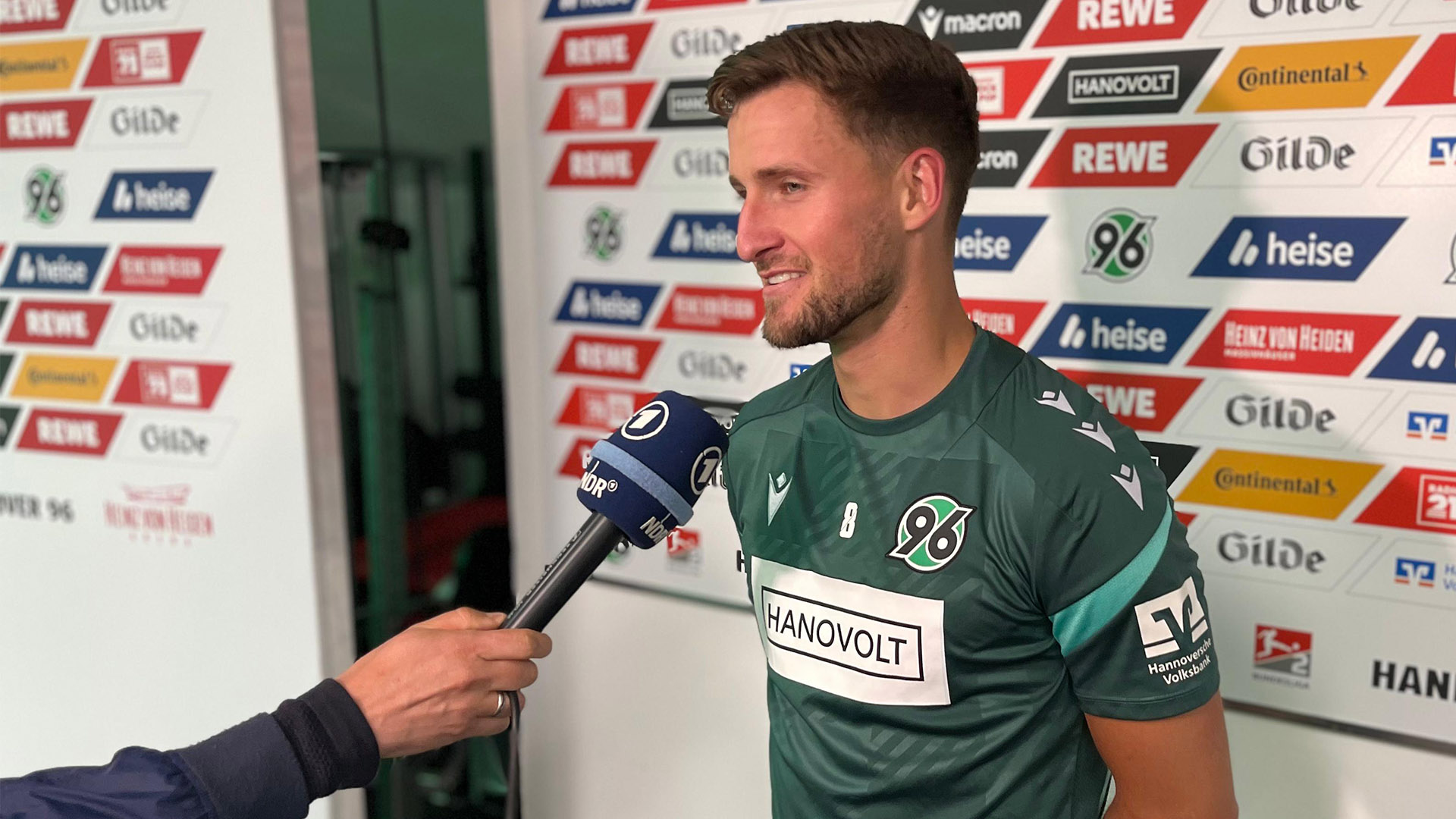 Enzo Leopold spricht am NDR-Mikrofon, im Hintergrund ist eine Stellwand mit Sponsorenlogos. 