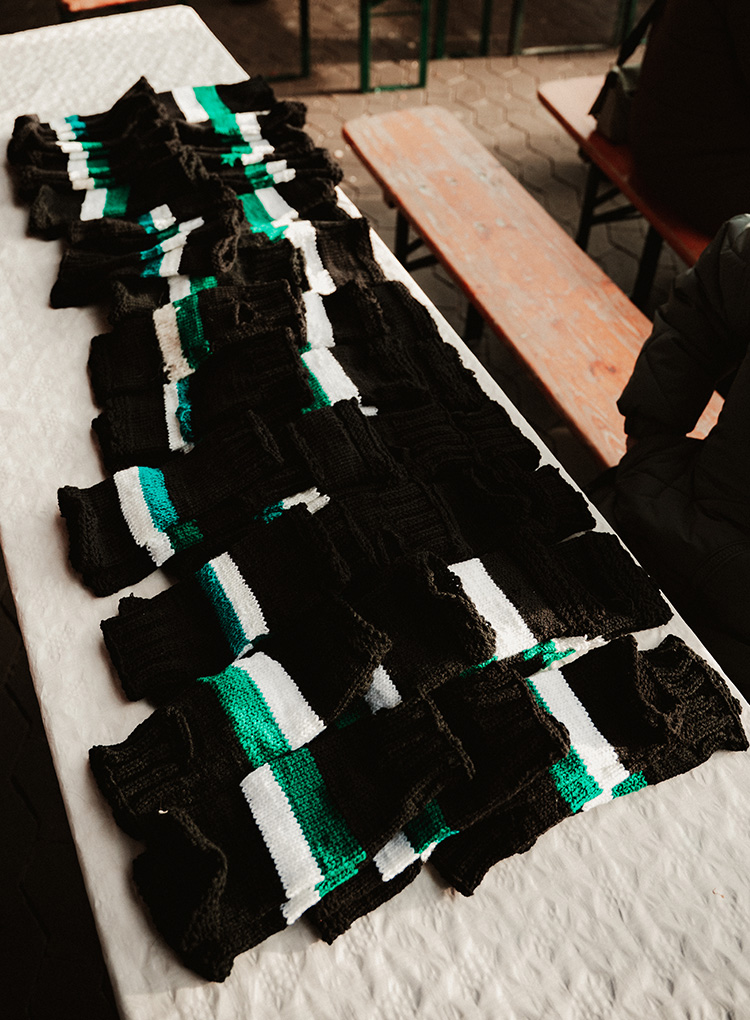 Auf einem Tisch liegen viele schwarze gestrickte Stulpen ordentlich nebeneinander. Jede Stulpe hat einen grünen und weißen Streifen.