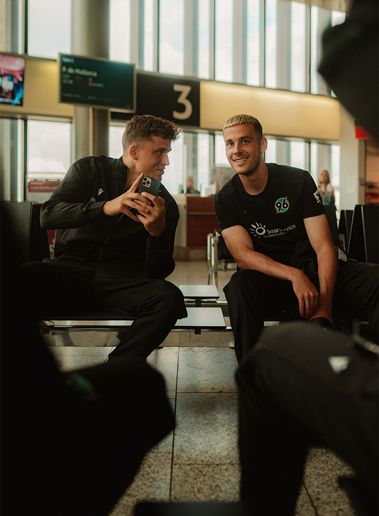 Benedikt Pichler und Bastian Allgeier sitzen in der Wartehalle, Pichler zeigt Allgeier etwas auf dem Handy, Allgeier lacht in die Kamera.
