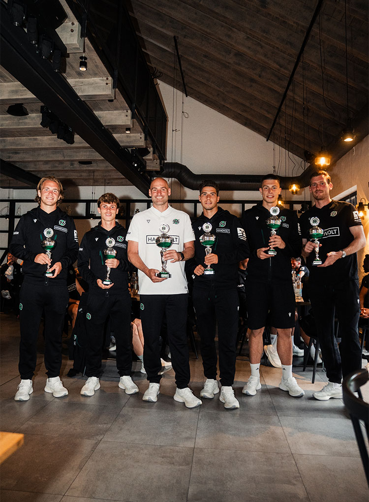 Fünf Spieler in dunklen Trainingssachen und ein Trainer in weißen Sachen posieren mit Pokalen.
