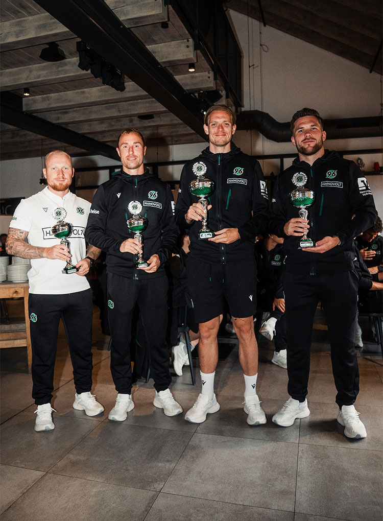 Drei Spieler in dunklen Trainingssachen und ein Trainer in weißen Sachen posieren mit Pokalen.