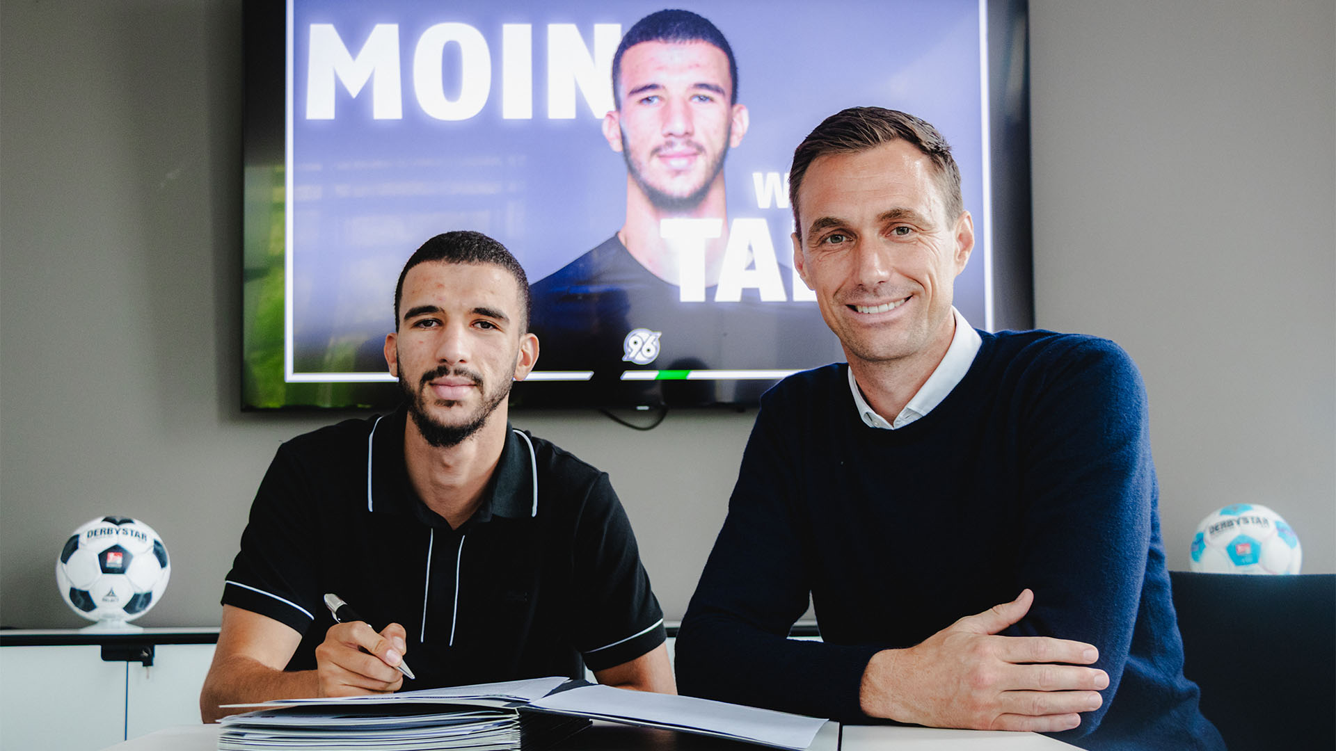 Hannover-96-Neuzugang Waniss Taibi und 96-Geschäftsführer Marcus Mann sitzen bei der Vertragsunterschrift in Manns Büro.