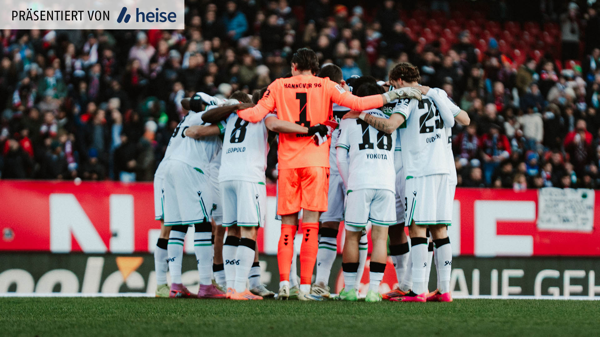 Die Startelf von Hannover 96 steht im Kreis, mit den Armen umeinander und leicht nach vorne gebeugt. Die Feldspieler tragen weiße Trikots.