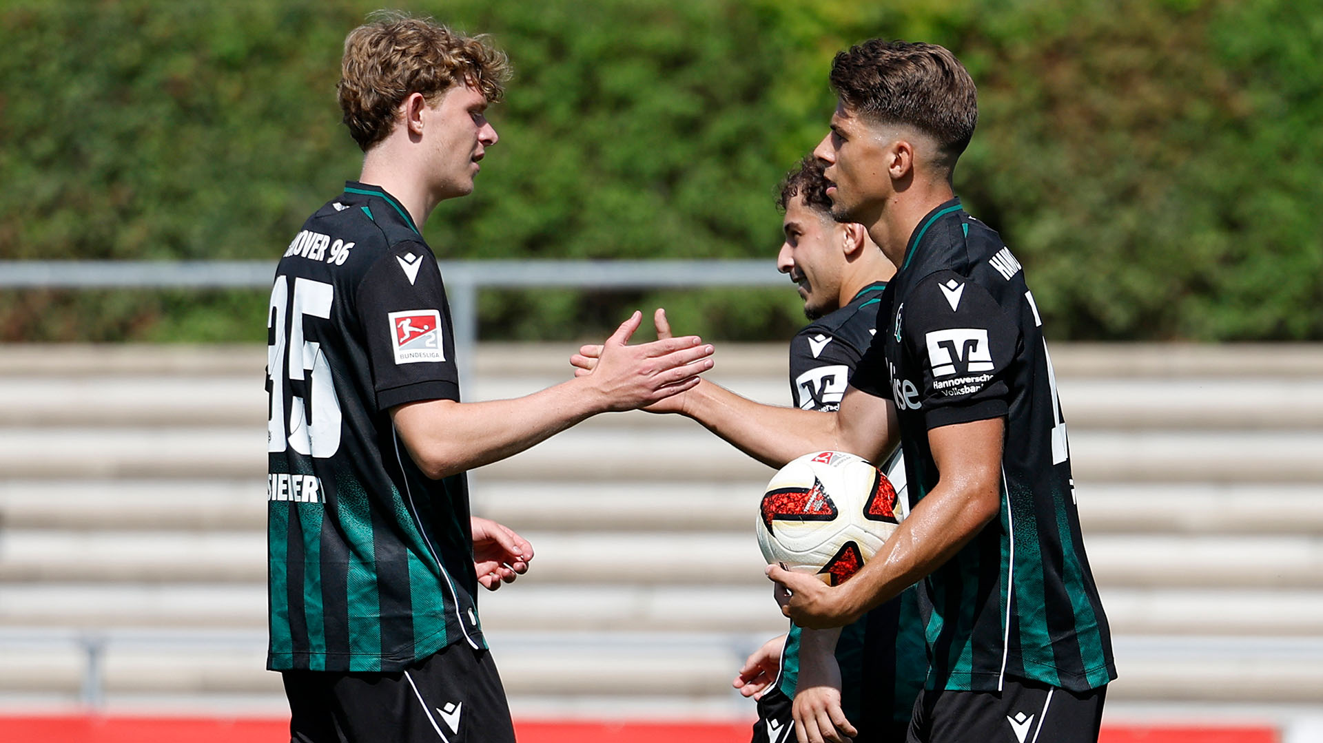 Zwei 96-Spieler in Auswärtstrikots klatschen miteinander ab, im Hintergrund ist ein weiterer Spieler zu sehen.