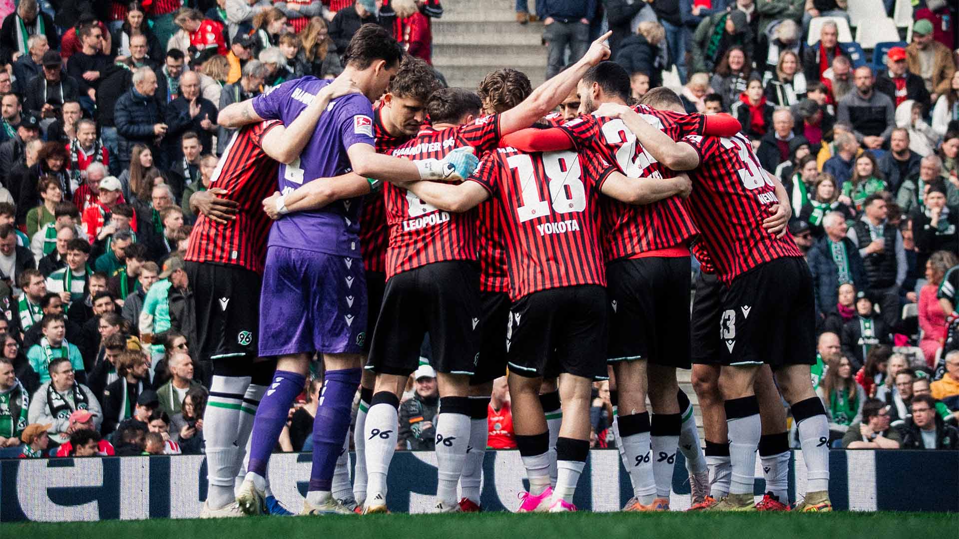 Die 96-Mannschaft bildet einen Kreis in Heimtrikot, im Hintergrund Fans auf den Tribünen.