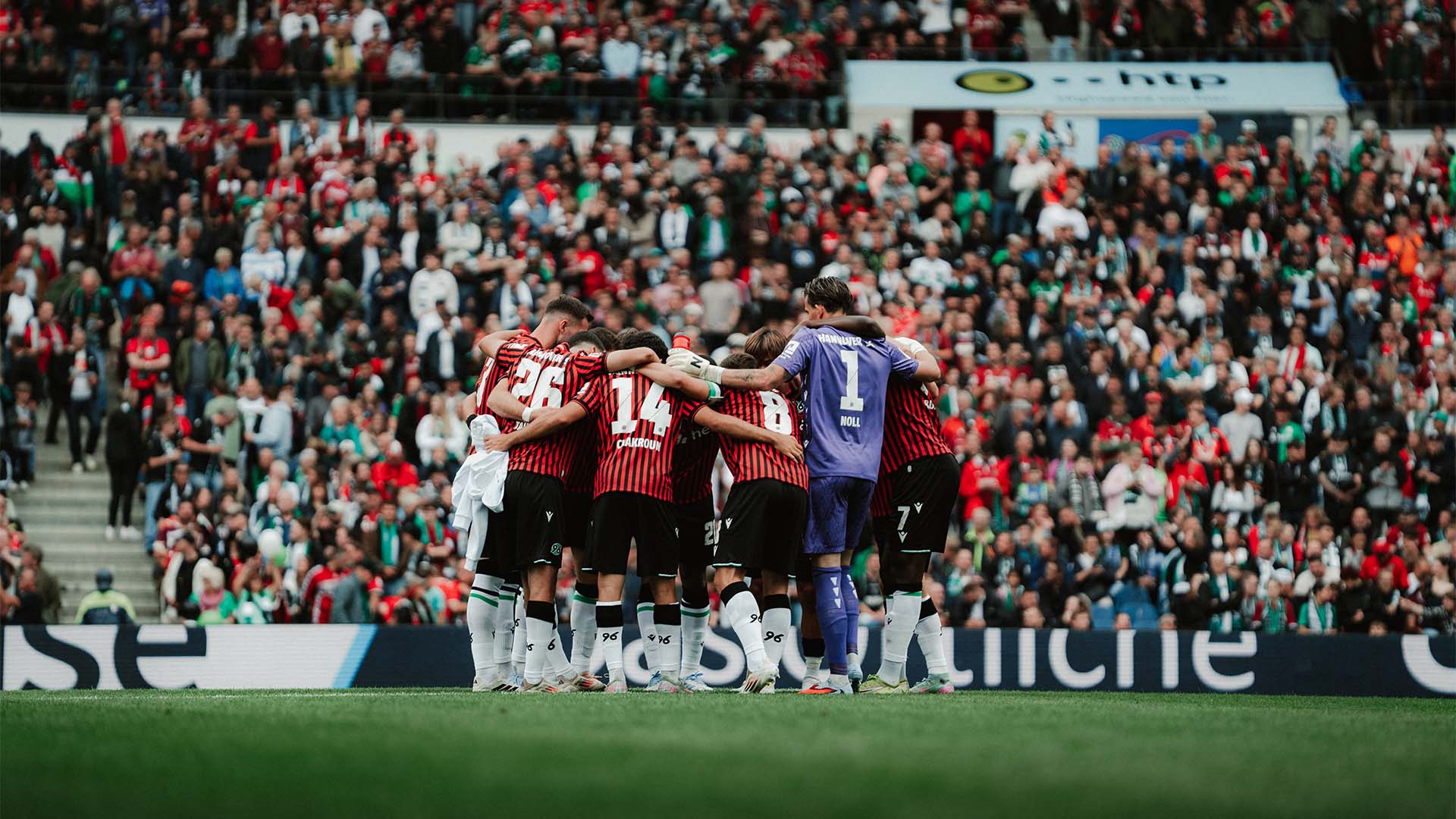 Die 96-Mannschaft bildet vor dem Spiel gegen Kaiserslautern einen Kreis, im Hintergrund sind volle Ränge zu sehen.