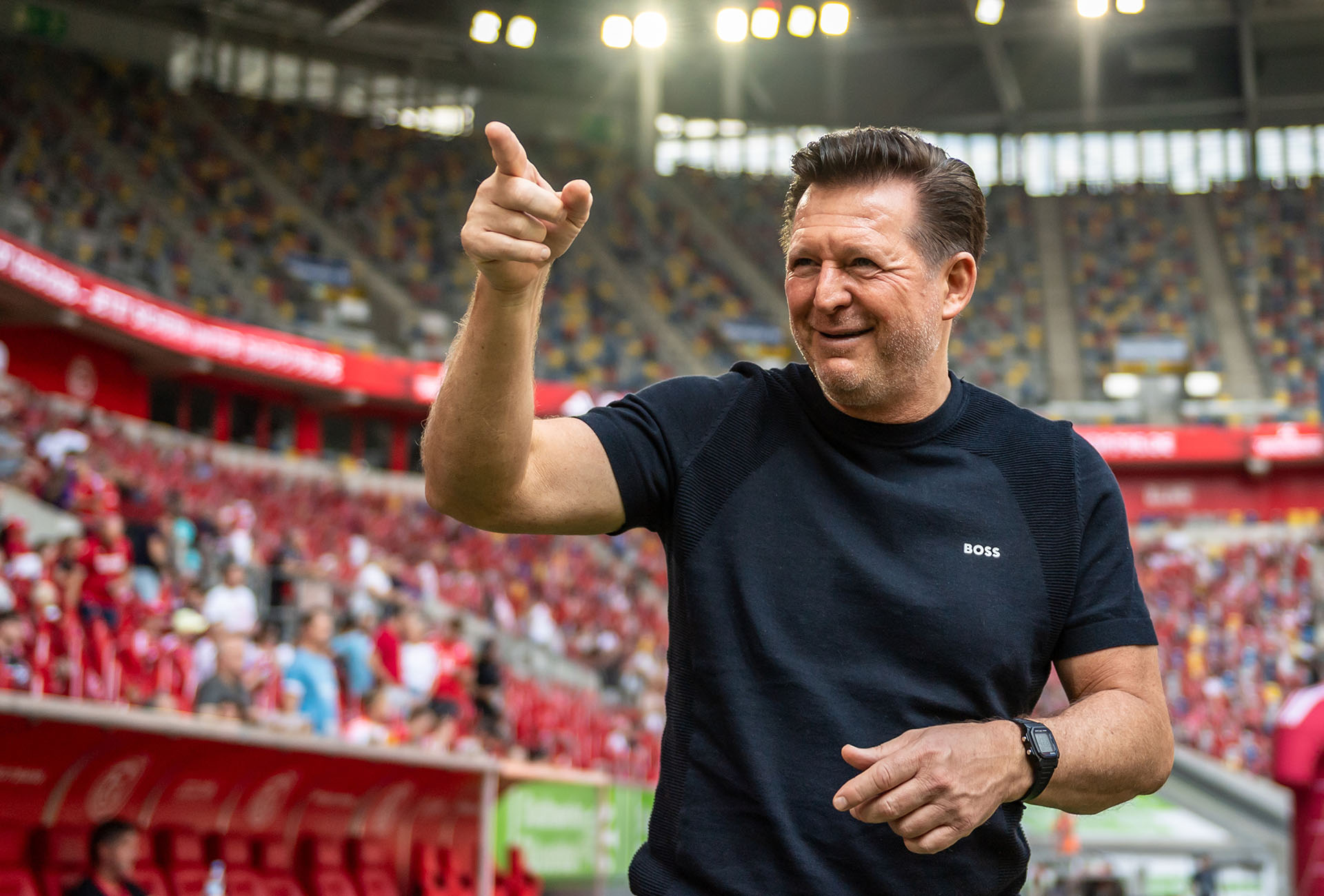 Christian Titz im Düsseldorfer Stadion, lächelt und zeigt mit dem rechten Zeigefinger nach oben.