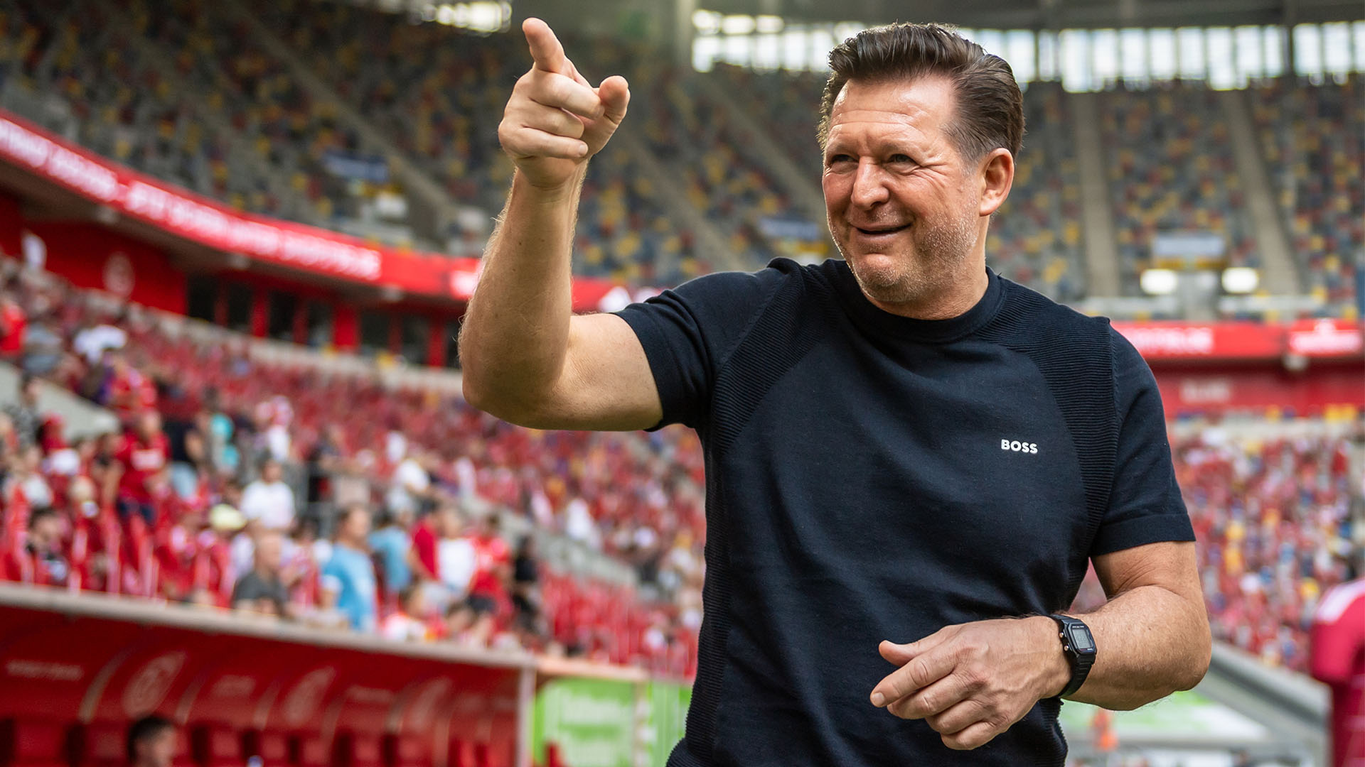 Christian Titz im Düsseldorfer Stadion, lächelt und zeigt mit dem rechten Zeigefinger nach oben.