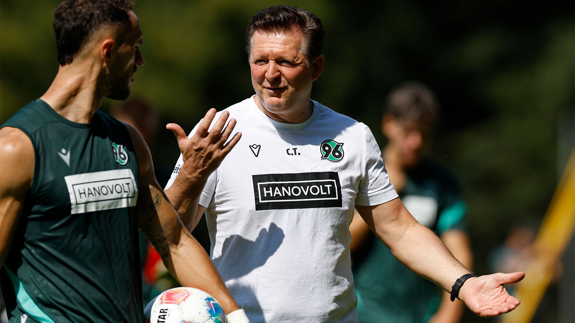 Christian Titz gestikuliert, spricht und lächelt dabei leicht, Virgil Ghita mit Ball in der Hand dreht sich zu ihm um.  