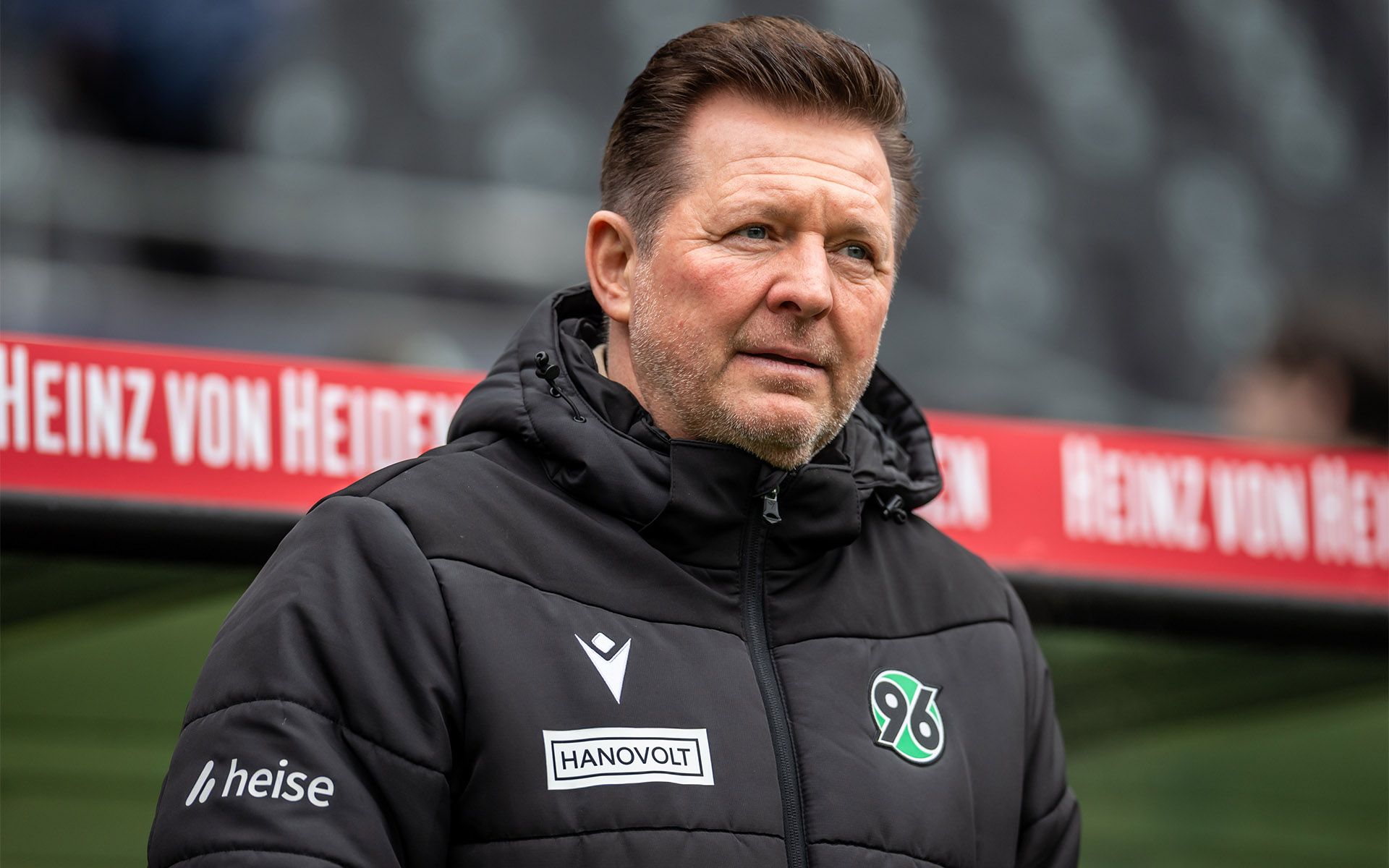 Christian Titz in schwarzer 96-Jacke vor der Trainerbank im Stadion.