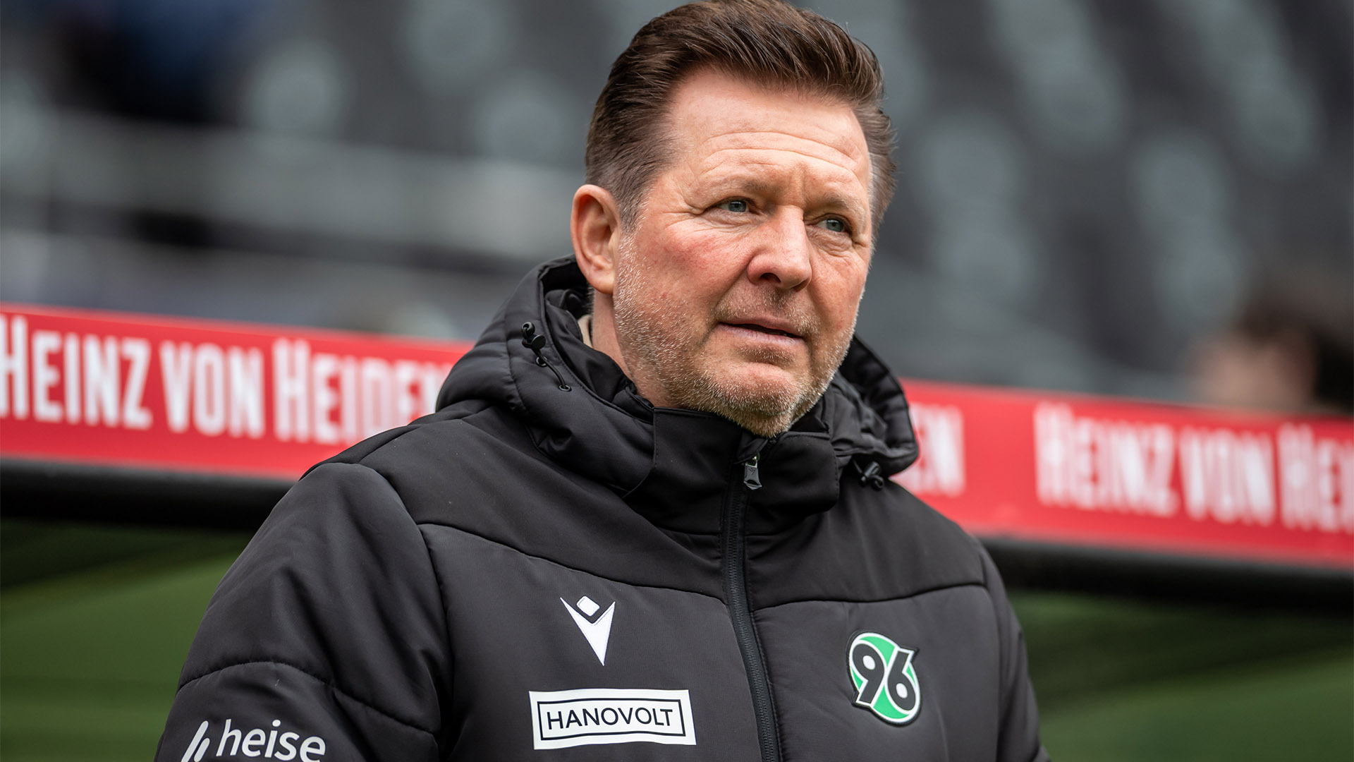 Christian Titz in schwarzer 96-Jacke vor der Trainerbank im Stadion.