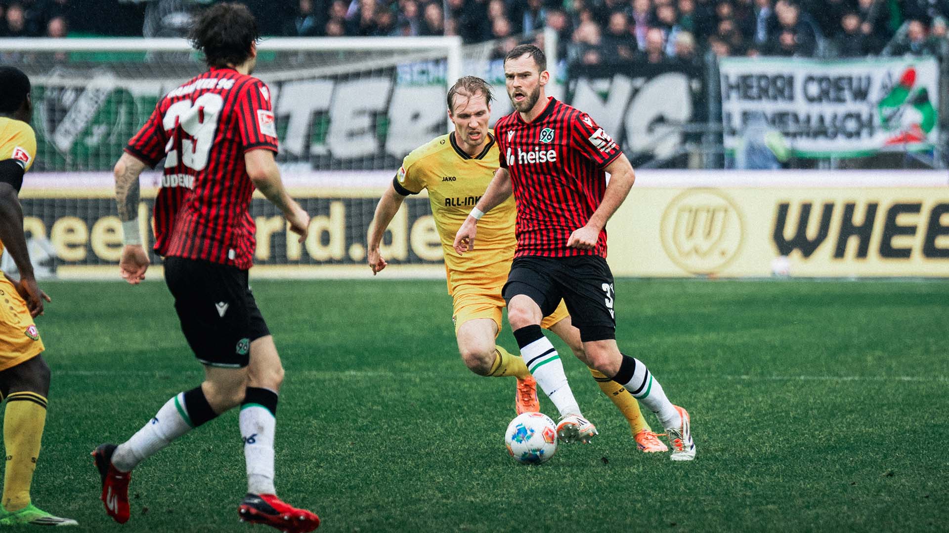 Boris Tomiak im rot-schwarz gestreiften Trikot führt den Ball mit dem rechten Außenrist und schaut nach vorn, während ein Gegenspieler im gelben Trikot dicht hinter ihm herläuft.