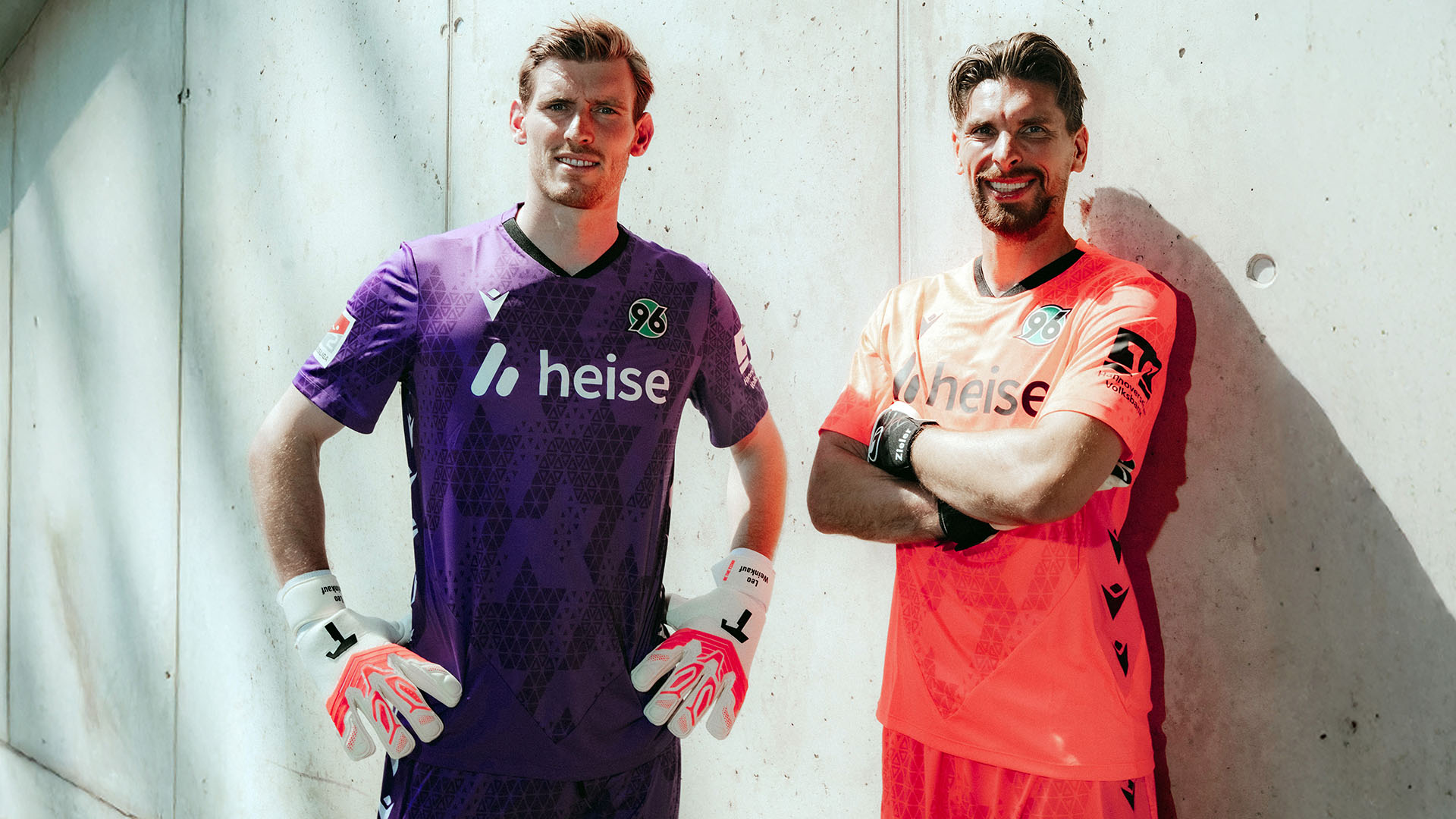 Leo Weinkauf im lilafarbenen Torwarttrikot und Ron-Robert Zieler im korallfarbenen Torwarttrikot posieren vor einer Wand.  