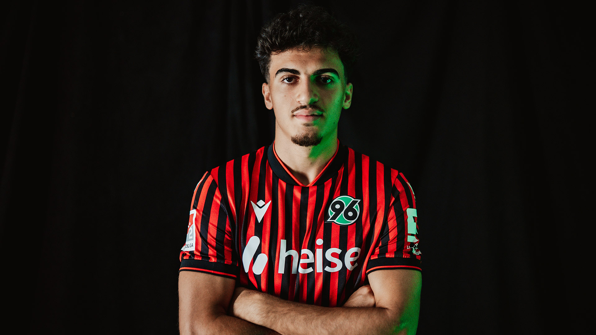 Husseyn Chakroun mit verschränkten Armen im Heimtrikot vor schwarzem Hintergrund.