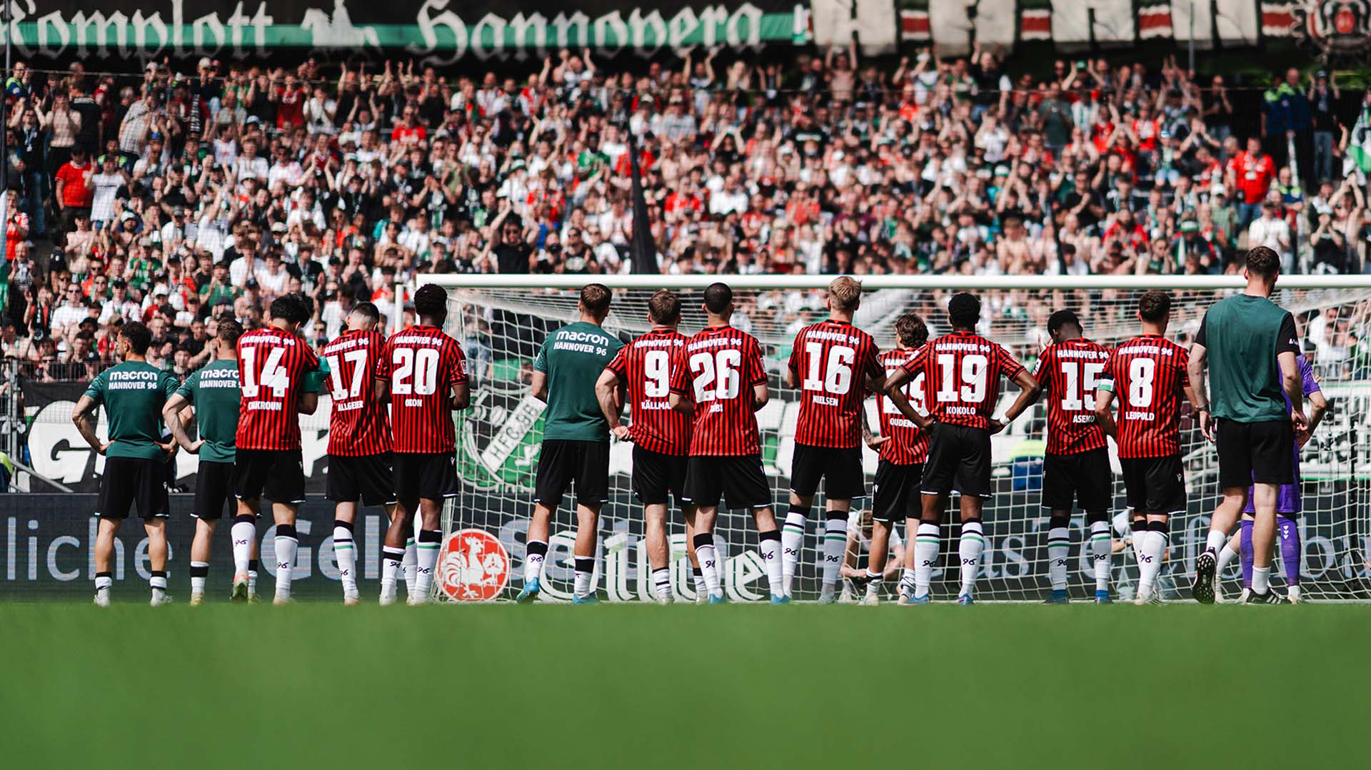 Ein Teil der 96-Spieler steht in Heimtrikots vor der Fankurve in der Heinz von Heiden Arena, die Spieler sind von hinten zu sehen.