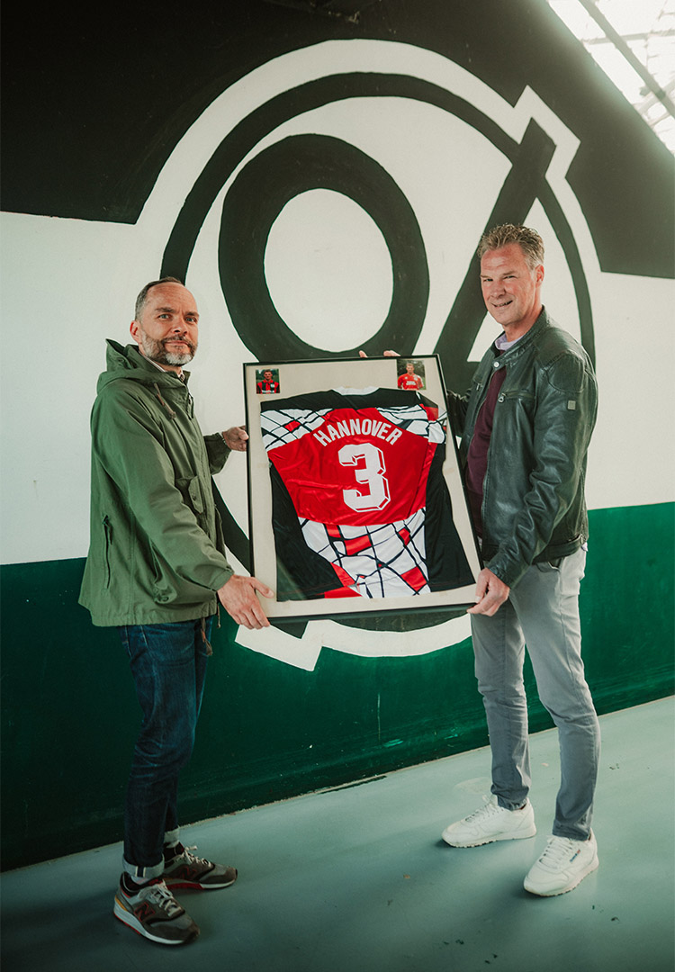 Jannis Busse (links) und Jörg-Uwe Klütz halten den Rahmen mit dem Pokalsiegertrikot, im Hintergrund eine 96 im Norden des Stadions.