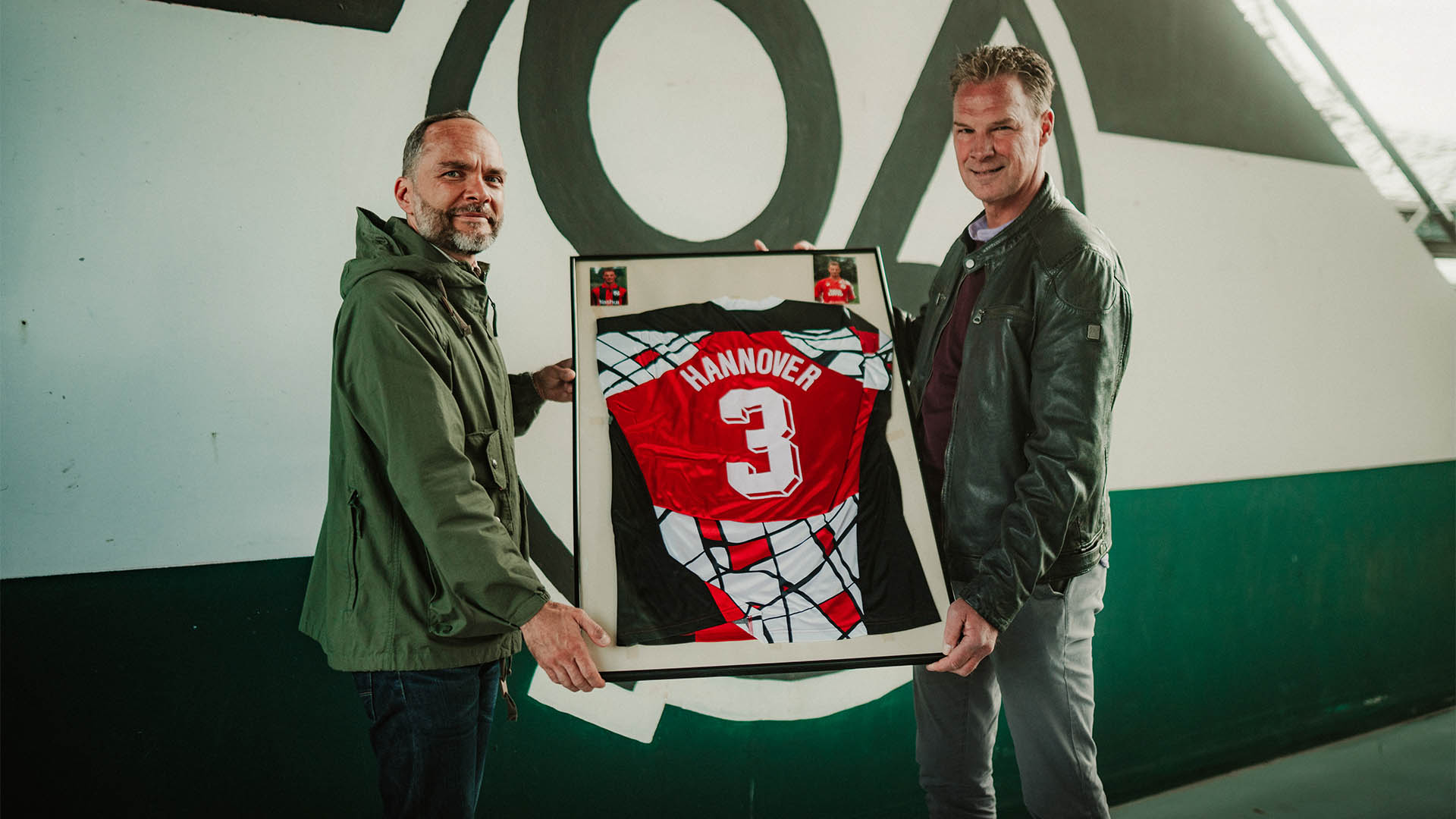 Jannis Busse (links) und Jörg-Uwe Klütz halten den Rahmen mit dem Pokalsiegertrikot, im Hintergrund eine 96 im Norden des Stadions.
